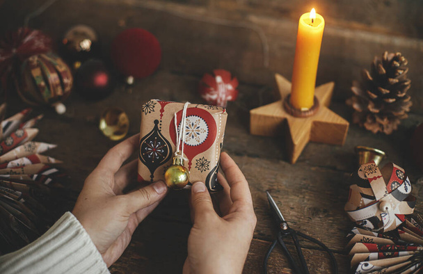 Ręce trzymając pudełko zapakowane w świąteczny czerwony papier na rustykalnym drewnianym tle stołu ze świecą, nożyczki, ozdoby. Atmosferyczny obraz. Wesołych Świąt i Wesołych Świąt! - Zdjęcie, obraz