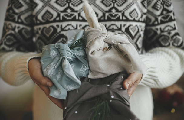 Zero waste and eco friendly presents, Christmas Furoshiki gifts. Woman hands in cozy sweater holding wrapped xmas gift in fabric in scandinavian room. Atmospheric moody image, nordic style. - Photo, image