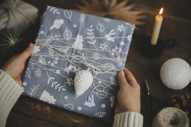 Hands holding stylish christmas gift wrapped in fabric on rustic wooden table with scissors, craft paper star, candle. Atmospheric moody time, nordic style. Happy Holidays! - Фото, зображення