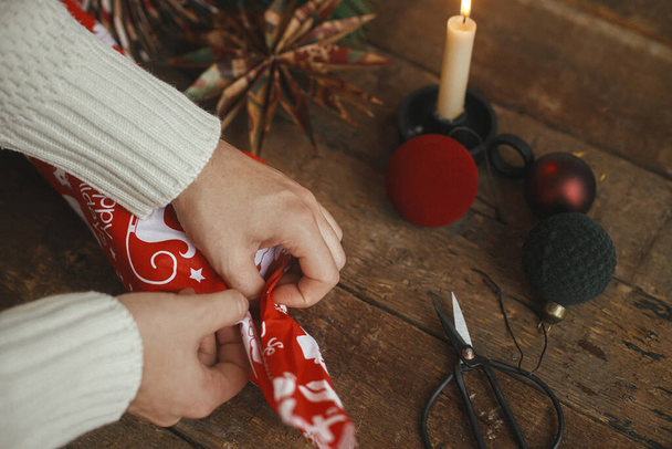 Noel hediyesini kırmızı şenlikli kumaşla saran eller kırsal ahşap masa üzerinde makasla, kağıt yıldızıyla, mumla, süslerle. Atmosferik karamsar zaman. Sıfır ziyan bayramı, furoshiki Noel hediyesi - Fotoğraf, Görsel