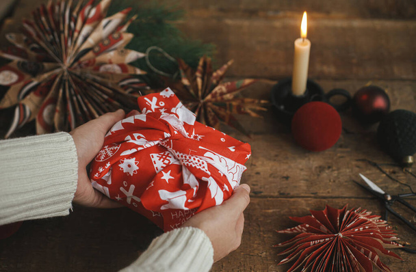 Χέρια κρατώντας χριστουγεννιάτικο δώρο τυλιγμένο σε κόκκινο εορταστικό ύφασμα σε ρουστίκ ξύλινο τραπέζι με ψαλίδι, αστέρι χαρτί, κερί, στολίδια. Ατμοσφαιρική κυκλοθυμική ώρα. Χριστουγεννιάτικο δώρο Furoshiki, διακοπές μηδενικών αποβλήτων - Φωτογραφία, εικόνα