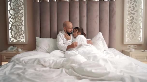 Jóképű fiatal férfi és lány fekszenek fehér köpenyben az ágyban gyengéden simogatják egymást.  - Felvétel, videó
