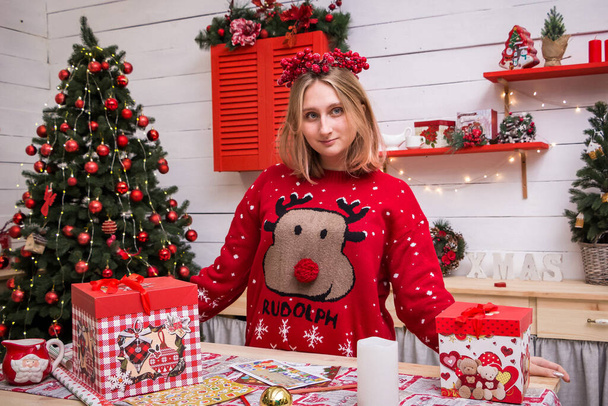 дівчина в вінку і новорічний червоний светр з подарунком і листом на Новий рік біля ялинки на різдвяній кухні
 - Фото, зображення