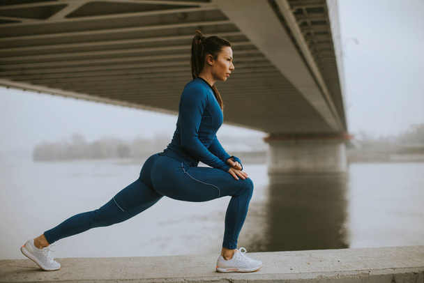 Pretty young woman in blue track suit stretching before workout by the river at autumn morning - Foto, immagini