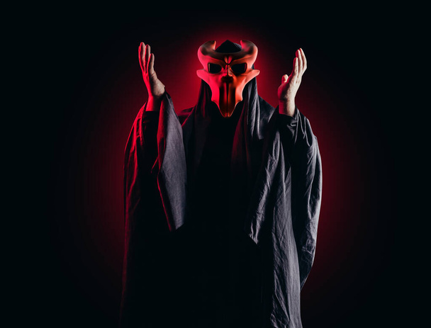 Страшный ужас оккультный священник секты в черном капюшоне и металлической маске на черном фоне с красным свечением. - Фото, изображение