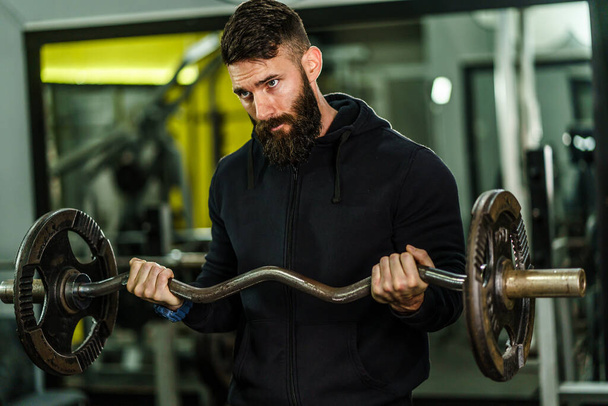 Front view portrait of one young caucasian man male athlete bodybuilder training at the gym workout using barbell biceps curls wearing black hoodie dark hair and beard standing weight lifting - Foto, imagen