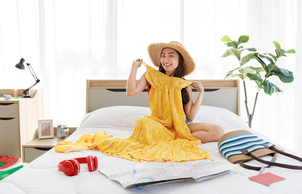 Asian young happy female traveler wears big hat laughing smiling sitting trying on yellow summer long dress on bed preparing personal stuff for sea and beach dream destination vacation holiday trip. - Photo, Image