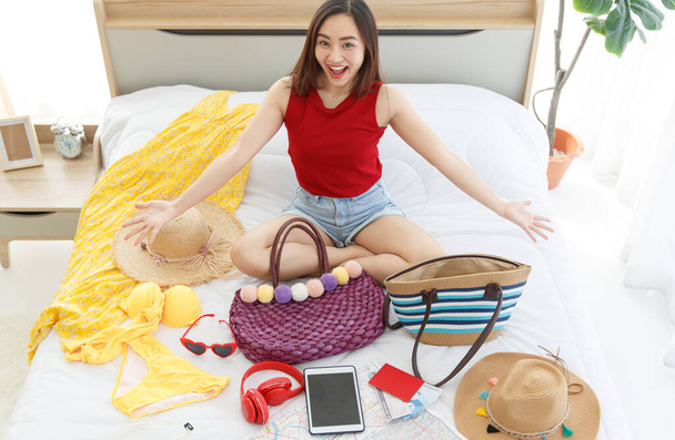 Asian beautiful young happy female traveler laughing smiling look at camera sitting showing preparing personal summer traveling stuff on bed in bedroom for weekend holiday vacation tourism trip. - Photo, Image