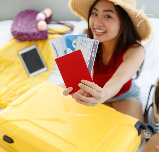 Red passport with air flight tickets in hands of Asian young female traveler wears big hat sitting look at camera packing personal traveling stuff to trolley luggage on bed in blurred background. - Foto, afbeelding