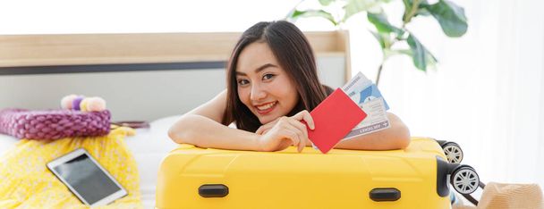 Asiatische junge glückliche Reisende mit rotem Reisepass und Flugtickets lehnen sich an großen gelben Trolley auf dem Bett und schauen in die Kamera, während sie persönliche Reisesachen für den Sommerurlaub einpacken. - Foto, Bild