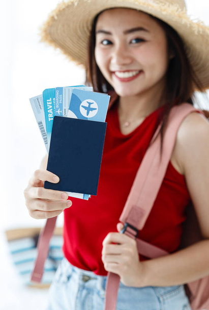 Nahaufnahme von blauem Reisepass und Flugtickets in der Hand einer glücklichen Reisenden aus Asien, die einen großen Hut und einen rosafarbenen Rucksack trägt und lächelnd in die Kamera blickt.. - Foto, Bild