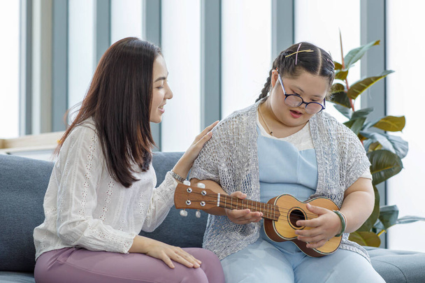 Mujer asiática enseñando a una joven con síndrome de Down a tocar el instrumento musical Ukulele en un sofá en la sala de estar en casa. - Foto, imagen