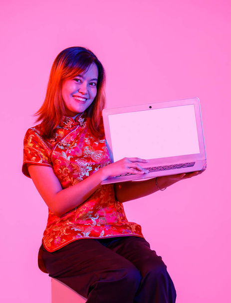 Portrait studio shot millennial Asian female model in red Chinese traditional cheongsam qipao shirt sitting holding showing presenting blank screen laptop computer product on white background. - Foto, afbeelding