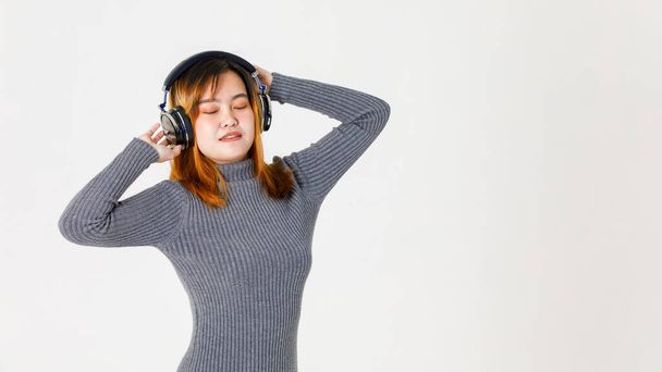 Studio shot van millennial gelukkig aziatisch jong geverfd haar vrouwelijk model in grijs coltrui gesloten ogen staan luisteren naar muziek via hoofdtelefoon headset dansen opgeheven handen op witte achtergrond. - Foto, afbeelding
