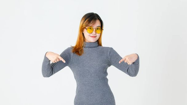 Ritratto studio girato di asiatica giovane modello femminile in maglione dolcevita grigio e occhiali da sole lente arancione in piedi puntando le dita indice verso il basso presentando promozione mostrando prodotto su sfondo bianco. - Foto, immagini