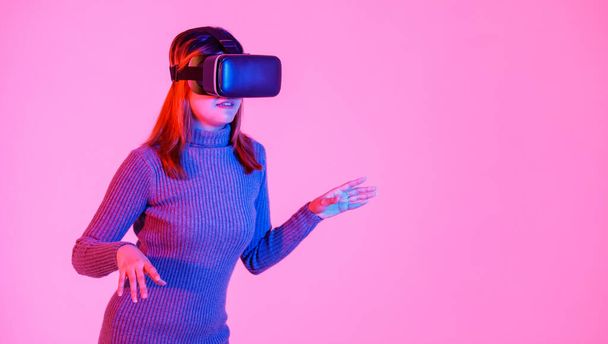 Studioaufnahme von asiatischen jungen Teenager weibliches Modell in grauem Rollkragenkleid trägt künstliche Intelligenz Virtual Reality vr Videospielbrille Headset spielt 3d-Konsole Gameplay auf rosa Licht Hintergrund. - Foto, Bild