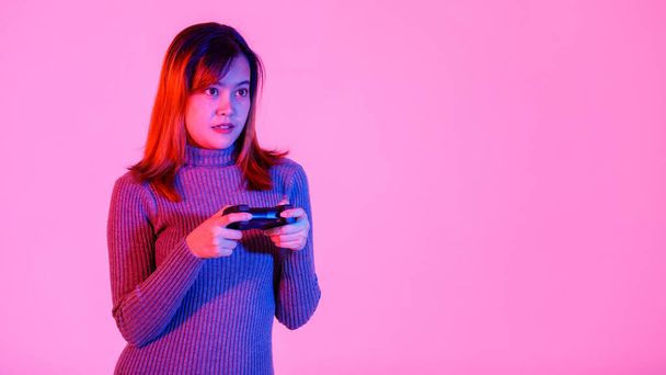 Portrait closeup studio shot of Asian young teen female model gamer in gray turtleneck dress standing smiling holding 3d video game dualshock console joystick in hands playing in pink light and background. - 写真・画像
