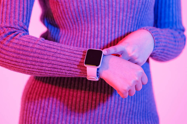 Close up studio shot of digital black blank empty screen touchscreen smartwatch trendy gadget on woman wrist who in gray long sleeve sweater dress pointing with index finger on pink light background. - Photo, Image