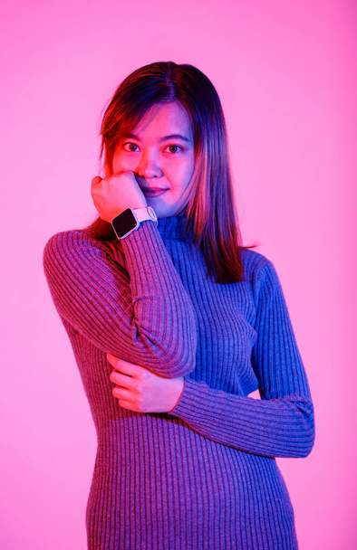 Portret studio strzał azjatycki miejski modny kobieta model w szary golf sweter stojący uśmiechnięty spojrzenie na aparat skrzyżowane ramię pokazując pusty ekran smartwatch gadżet na różowym tle światła. - Zdjęcie, obraz