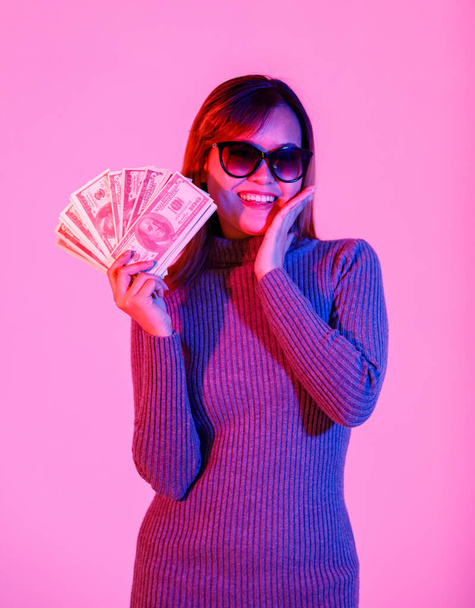 Retrato de estudio de tiro de Asiático joven rico modelo femenino fresco en suéter de cuello alto gris y gafas de sol de lentes negras que contienen billetes de cien dólares pila de moneda sobre fondo rosa claro. - Foto, imagen
