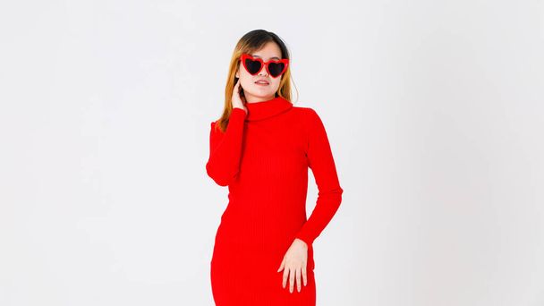 アジアの自信に満ちた女性ファッションモデルのポートレートスタジオショットカジュアルなファッショナブルな流行の赤の長袖タートルネックドレスでハート形の黒レンズサングラススタンドセクシーポージング上の白の背景. - 写真・画像