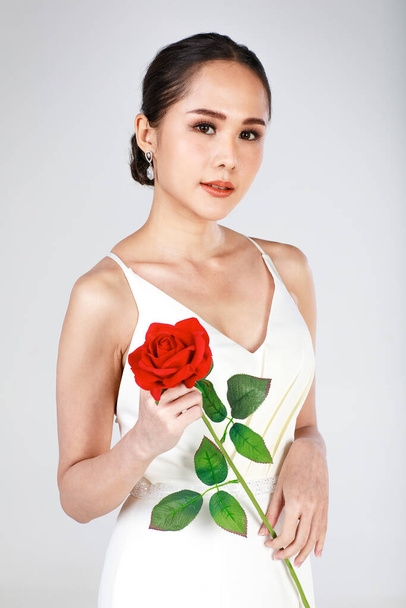 Портрет молодой привлекательной азиатки в белом свадебном платье с красной розой на белом фоне. Концепция предсвадебной фотографии. - Фото, изображение