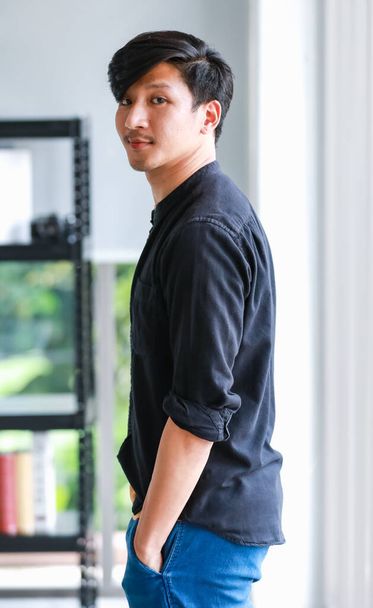 Портрет молодого веселого азиатского профессионального фотографа-мужчины, стоящего у окна в современном офисе, позирующего, держа ретро-камеру и улыбаясь - Фото, изображение