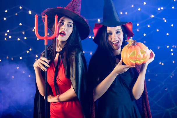 Studio shot of Asian Caucasian heureux modèles féminins sexy en robe de sorcière rouge noire costume grand chapeau et châle posant tenant trident et visage de citrouille dans la fête d'Halloween sur fond de fumée ampoule. - Photo, image