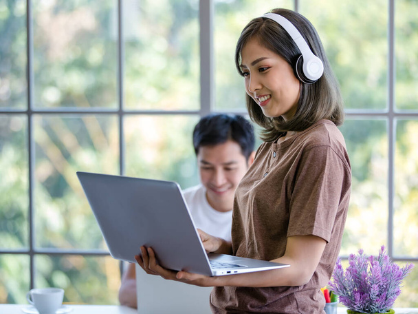 Пара молодых азиатских офисных работников, работающих над проектом, используют ноутбук и блокнот в домашнем офисе в удобной футболке. Пока леди слушает музыку в наушниках. - Фото, изображение