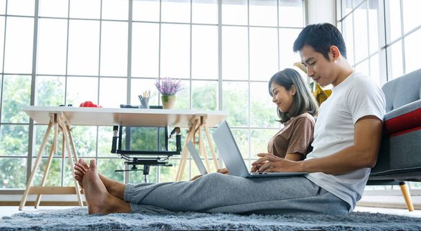 Jong Aziatisch koppel draagt comfortabel t-shirt en werkt thuis samen met een laptop. Conceptwerk van thuis uit. - Foto, afbeelding
