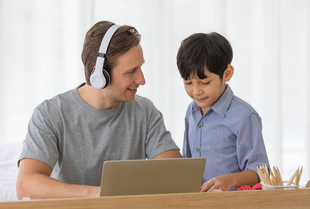 Beyaz bir baba, evde birlikte ders çalışırken küçük çocuğuna ev ödevlerinde dizüstü bilgisayar kullanarak yardım ediyor. Çocuk için çevrimiçi öğrenme kavramı. - Fotoğraf, Görsel
