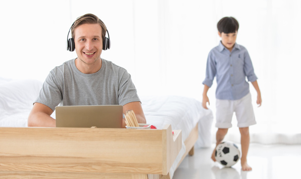 Νεαρός Καυκάσιος πατέρας που δουλεύει με το λάπτοπ του και φοράει ακουστικά στο σπίτι ενώ το παιδί του παίζει στο παρασκήνιο. Έννοια εργασία από το σπίτι. - Φωτογραφία, εικόνα