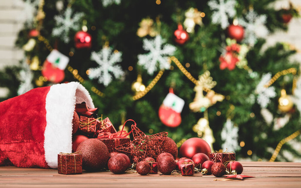 Κοντινό πλάνο των κόκκινων διακοσμητικών στοιχείων σφαίρα αστέρι παρόν κουτιά δώρων ρίχνει από το καπέλο Χριστουγέννων στο ξύλινο τραπέζι μπροστά από όμορφη διακόσμηση πλήρως με πάγο νιφάδες κάλτσες πεύκο δέντρο σε θολή φόντο. - Φωτογραφία, εικόνα
