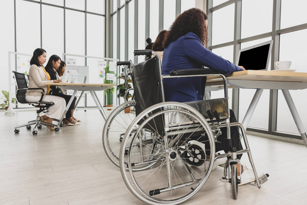 Aasialaiset liikenaiset, mukaan lukien vammainen nainen, joka istuu pyörätuolissa ja tekee kovasti töitä kannettavan tietokoneen ääressä toimistossa. Liiketoimintakokouksen käsite. - Valokuva, kuva
