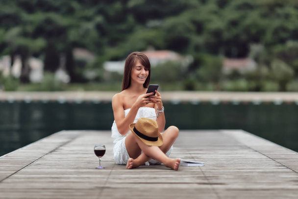 A Caucasian female model sitting on a wooden pier on a lake in Montenegro and using her mobile phone - Φωτογραφία, εικόνα