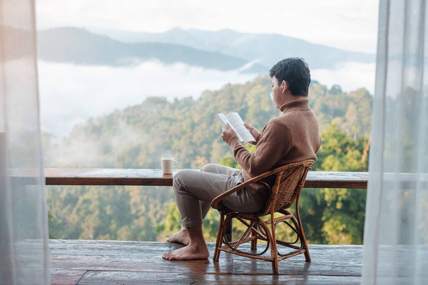 fiatalember könyvet olvas az ablak mellett, és nézi a hegyi kilátást a vidéki otthonban a reggeli napfelkelte. SoloTravel, utazás, utazás és pihentető koncepció - Fotó, kép