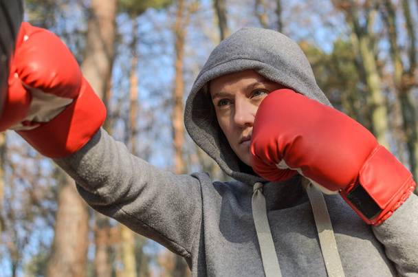 Young charming woman in red boxing gloves, dressed in a tracksuit with a hood, in a boxing training in the forest outdoors, beats a homemade punching bag from car tires - Фото, изображение