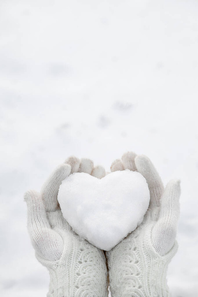 Μια καρδιά χιονιού στα χέρια φορώντας πλεκτά γάντια στο φόντο ενός καλύμματος χιονιού. - Φωτογραφία, εικόνα