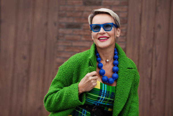Portrét krásné dospělé ženy se stylovými krátkými vlasy s krásným úsměvem a na sobě jasně zelený kabát, módní doplňky, modré sluneční brýle, korálky - Fotografie, Obrázek