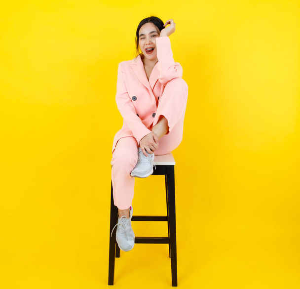 Симпатичная азиатка в розовом деловом костюме изображает шутливое лицо, привлекательно сидя на стульчике с локтем на коленях. Красивая портретная мода пастельного костюма, дизайн красоты для оживленной молодой женщины. - Фото, изображение