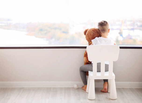 Widok z tyłu małego chłopca z pluszowym miśkiem siedzącym na krześle i wyglądającym przez okno na jesienny pejzaż miasta czekając na mamę w domu. Koncepcja dzieciństwa - Zdjęcie, obraz