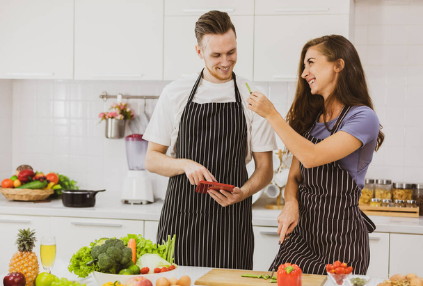 Delighted couple in aprons drinking hot beverages at counter with modern kitchenware in light kitchen with various products on table - Photo, Image