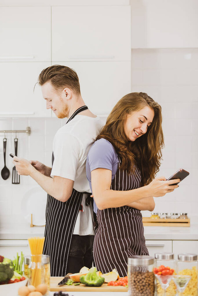 Encantada pareja en delantales tomando autorretrato en el teléfono inteligente cerca de la mesa con productos surtidos mientras cocinan juntos en la cocina ligera - Foto, imagen