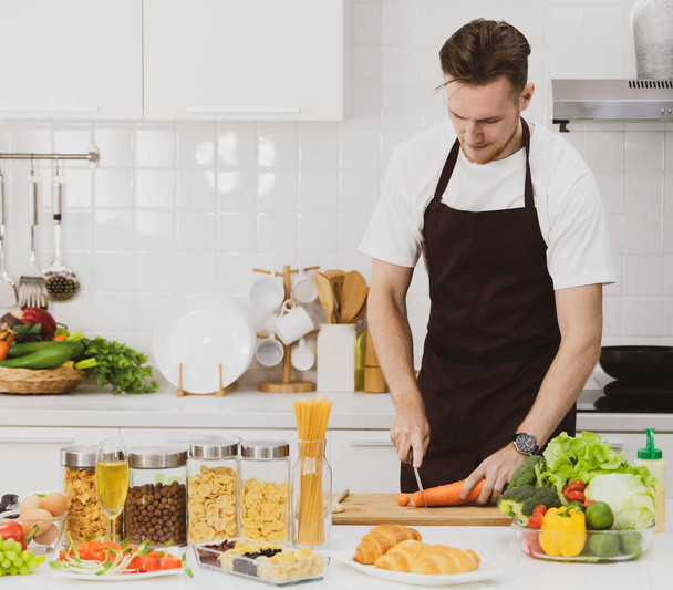 Fókuszált férfi szakács kötényben vágás nyers répa vágódeszkán, miközben áll az asztalnál válogatott termékek a modern konyhában - Fotó, kép