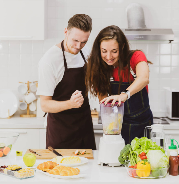Positief paar in schorten gelukkig samen tijdens het maken van verfrissende smoothie met blender aan tafel met diverse ingrediënten in licht moderne keuken - Foto, afbeelding