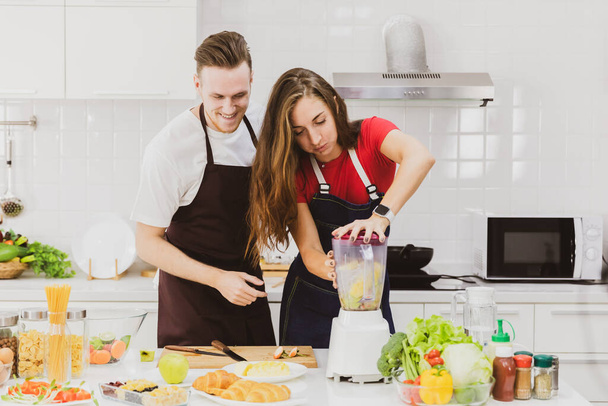Позитивна пара в фартухах щаслива разом під час приготування освіжаючої смузі з блендером за столом з різноманітними інгредієнтами в світлій сучасній кухні
 - Фото, зображення