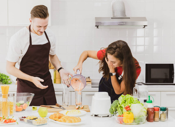 Positive couple in aprons pouring refreshing smoothie from blender into glass at table with assorted ingredients in light modern kitchen - Photo, Image