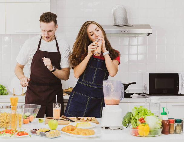 Pozitivní pár v zástěrách tanec s brýlemi u stolu s různými ingrediencemi a mixér při výrobě smoothie ve světle moderní kuchyně - Fotografie, Obrázek