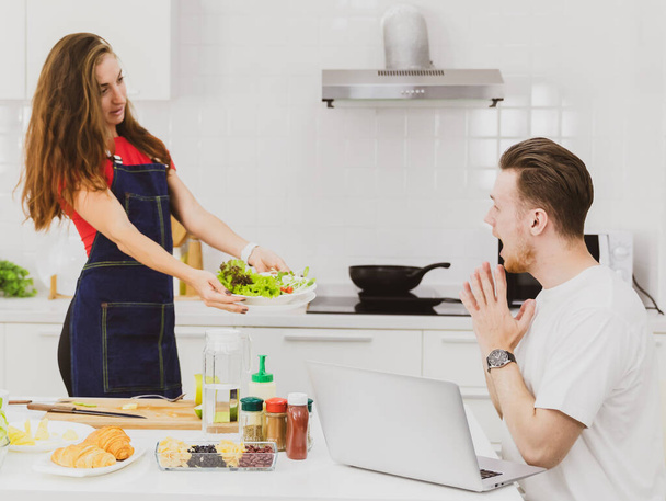 Vista lateral da namorada no avental mostrando tigela de salada fresca para namorado surpreso perto da mesa com produtos na cozinha - Foto, Imagem