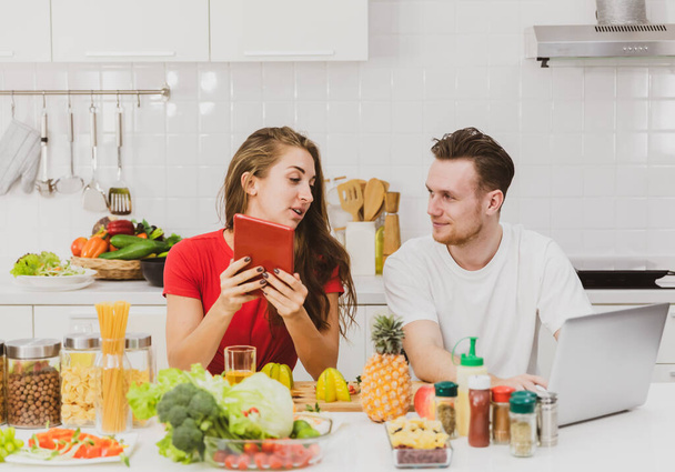 Coppia uomo e donna seduti insieme in cucina utilizzando tablet e laptop trovare cibo e formula di cottura via internet circondato da frutta e ingredienti. - Foto, immagini